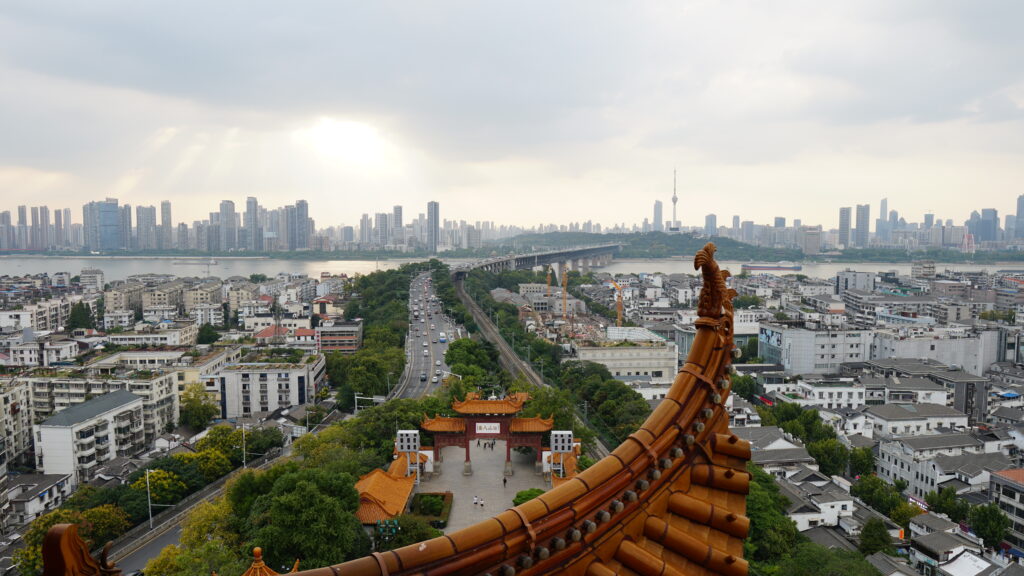 An aerial view of a cityscape with a river, tall buildings, a bridge, and traditional architecture in the foreground under a cloudy sky with sunlight peeking through showcases Wuhan's landmarks.