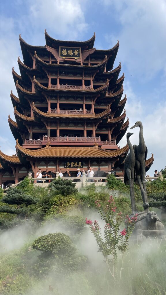 A tall, multi-tiered pagoda with intricate architectural details stands against a cloudy sky, with a garden featuring a crane statue and mist in the foreground. People can be seen on the pagoda's balconies, enjoying views of Wuhan's landmarks and sampling local food from nearby vendors.