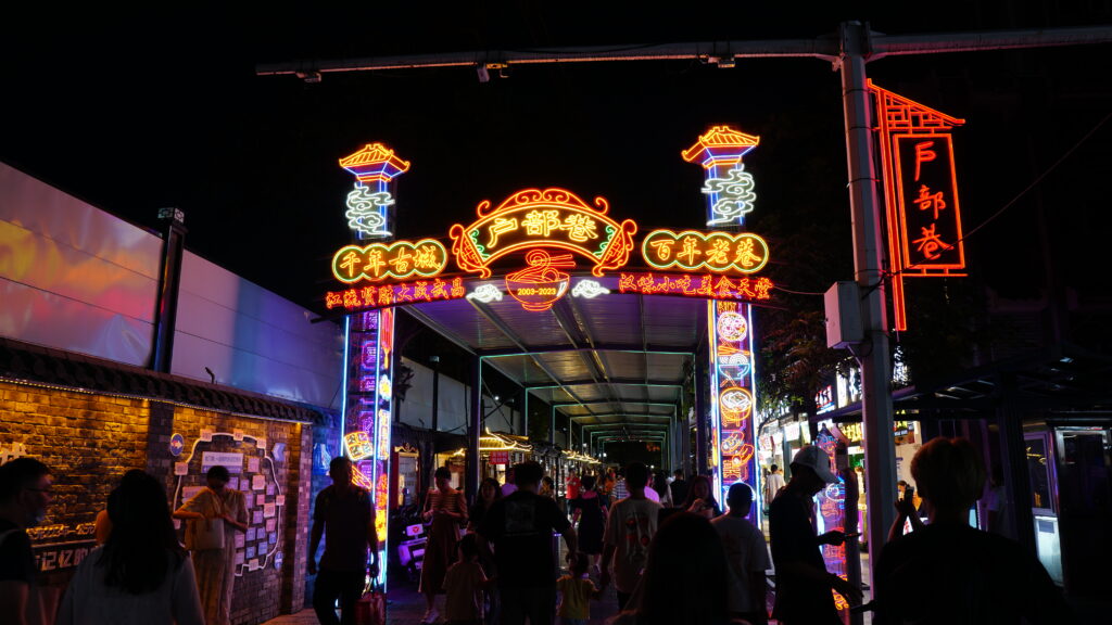 Pedestrians stroll through a bustling night market in Wuhan, illuminated by colorful lights and filled with food stalls and vendors.