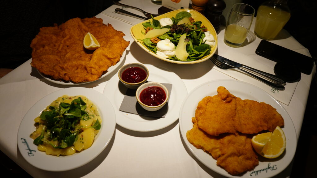 Image of plates of Wiener Schnitzel, salad and jam, with drinks, neatly arranged on a dining table for our Vienna travel and food good.