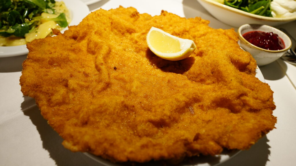 Image of a plate of Wiener Schnitzel on a table with a lemon, jam and some side dishes for our vienna travel and food good.