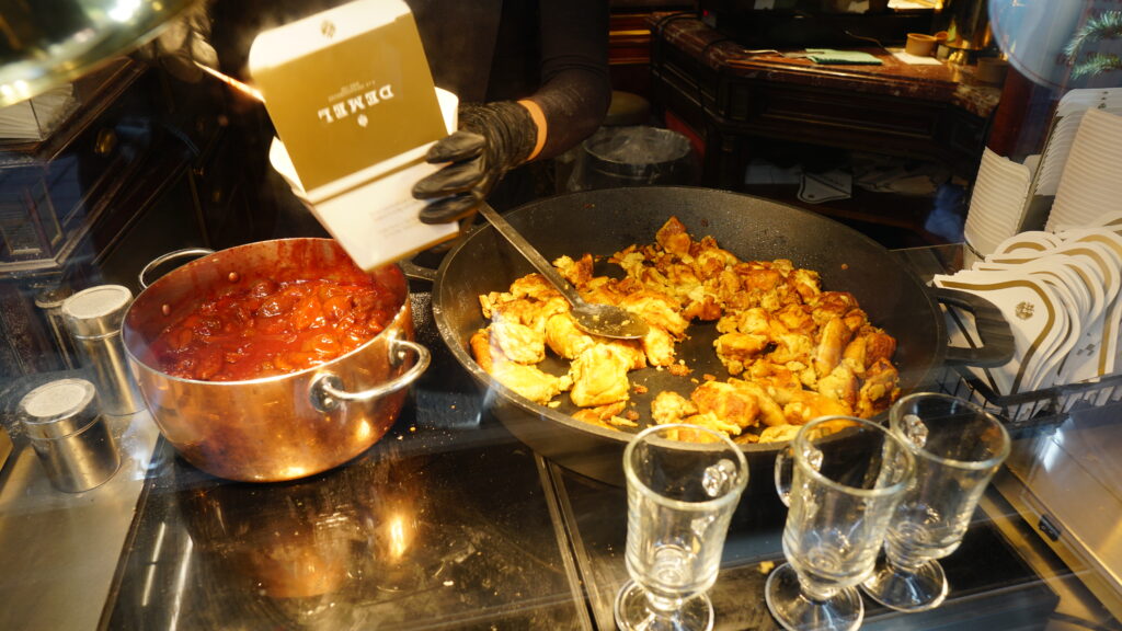 Kaiserschmarrn from a pan and jam from a pot are served in a paper box by a person wearing black gloves.