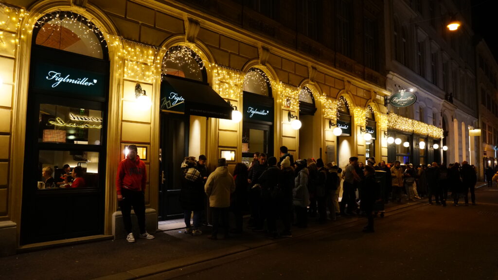 Customers lining up outside a restaurant in the evening, with a dark sky and illuminated signs.