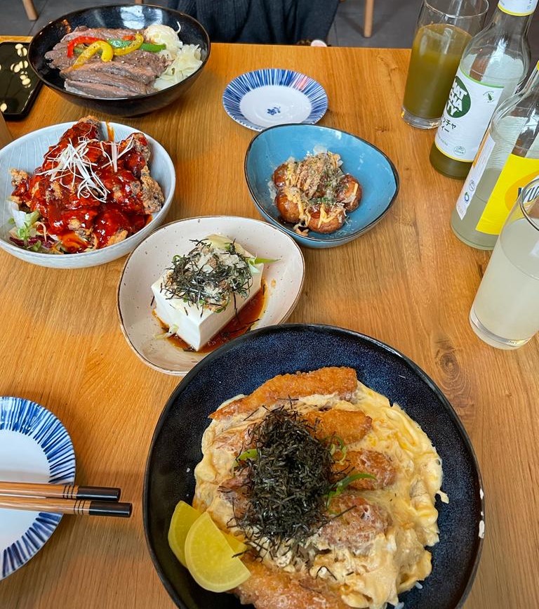 A table filled with various plates of food and drinks, including Japanese dishes such as steak don and katsudon.